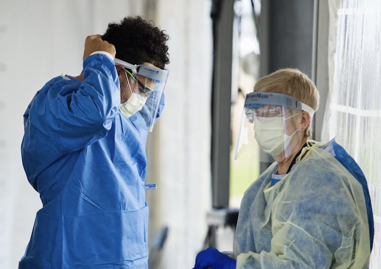 Two health-care professionals, one of whom is adjusting a face shield and the other is putting on a yellow protective gown.