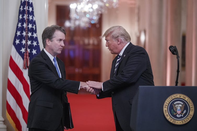 Donald Trump shakes hand of Brett Kavanaugh with US flag behind.