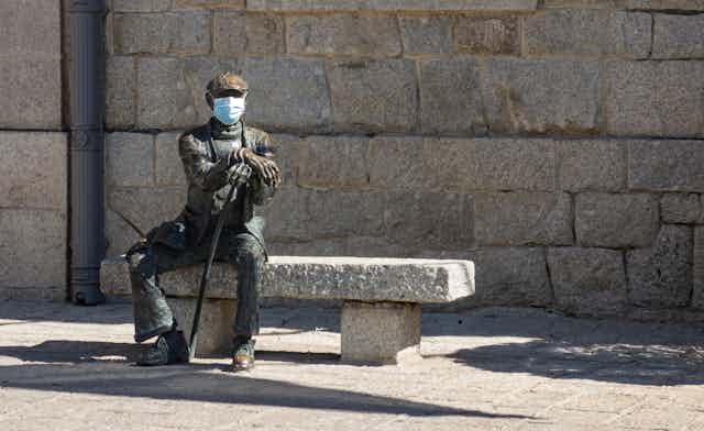 Estatua de un hombre sentado en un banco al que han colocado una mascarilla quirúrgica