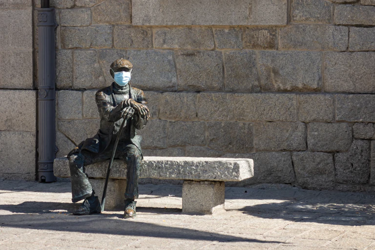 Estatua de un hombre sentado en un banco al que han colocado una mascarilla quirúrgica