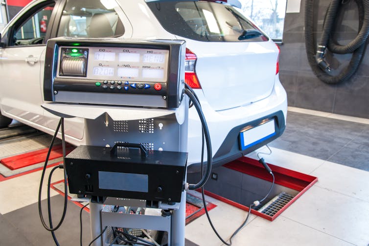 A passenger car in a testing facility with cables attached to the exhaust and a monitor displaying emissions data.