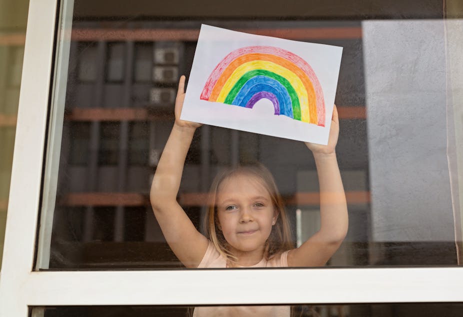 Little girl holding rainbow drawing up to the window.