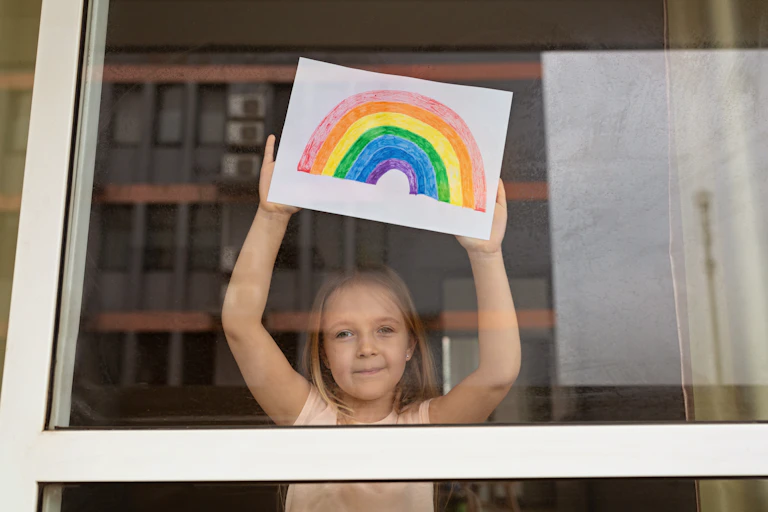 Little girl holding rainbow drawing up to the window.