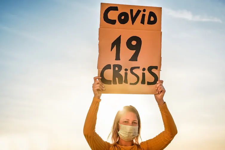 Una mujer con mascarilla sujeta un cartón con la leyenda: Covid 19 crisis.