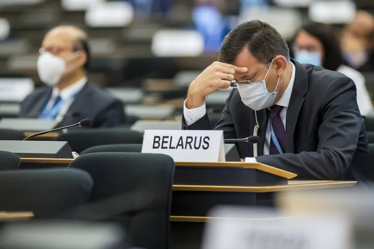 Man in maks holds head in hand in front of sign saying Belarus.