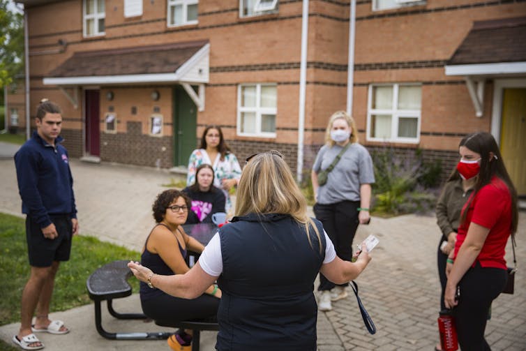 A person presents to a semi-circle of masked students standing outdoors.