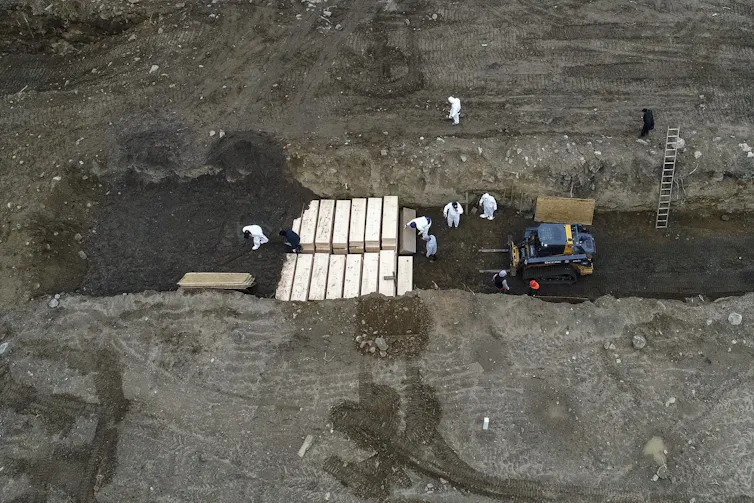 Workers wearing protective equipment place coffins in a trench.