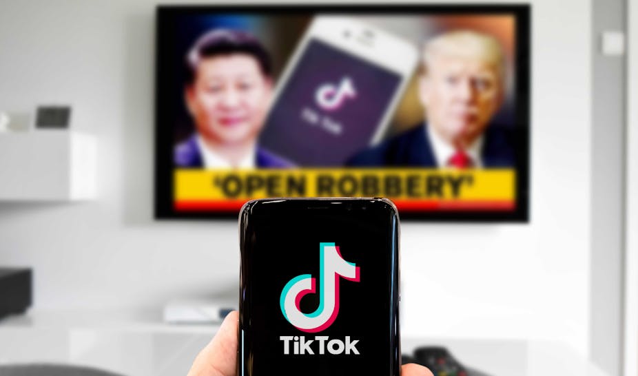 A person looks at TikTok on their phone while Donald Trump and Xi Jinping appear on a TV in the background.