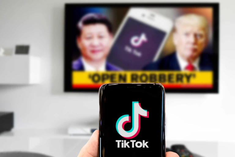 A person looks at TikTok on their phone while Donald Trump and Xi Jinping appear on a TV in the background.