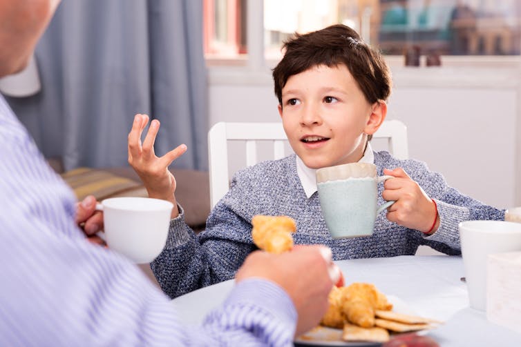 Boy talking to adult while drinking from a mug.