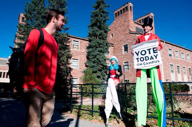 Man and woman on stilts in patriotic outfits hold a sign reading 'Vote Today on Campus' and talk to a student
