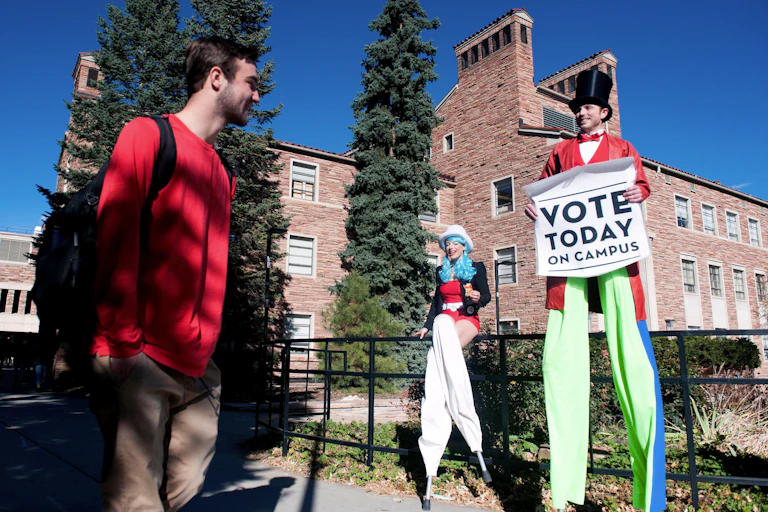 Man and woman on stilts in patriotic outfits hold a sign reading 'Vote Today on Campus' and talk to a student