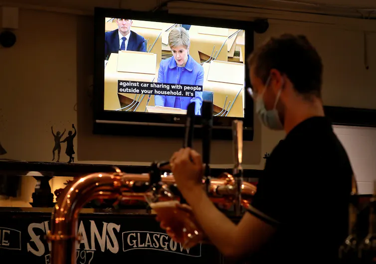 A man pulls pints in a pub while a television show Nicola Sturgeon addressing the Scottish parliament