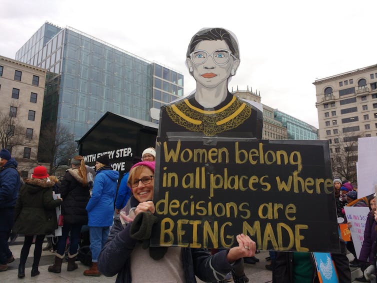 Woman in red hat at protest holding up a sign with an image of Ruth Bader Ginsburg and the text 'Women belong in all places where decisions are being made'