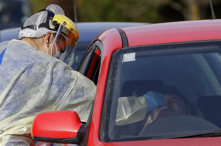 A medical person reaching into a car window to carry out a COVID-19 test on the driver.