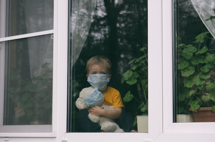 A child and a teddy, both wearing masks, look out a window.