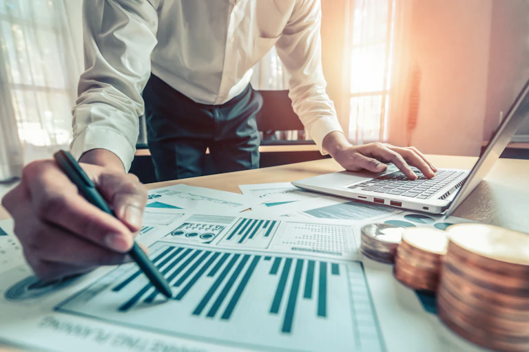 Formally dressed man looking over graph sheets on his desk with money stacked next to the sheets.