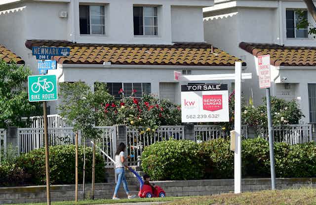 A for sale sign in front of a white California home, with a woman walking with her child on the sidewalk