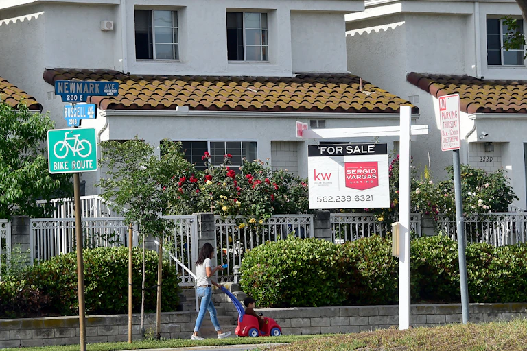 A for sale sign in front of a white California home, with a woman walking with her child on the sidewalk