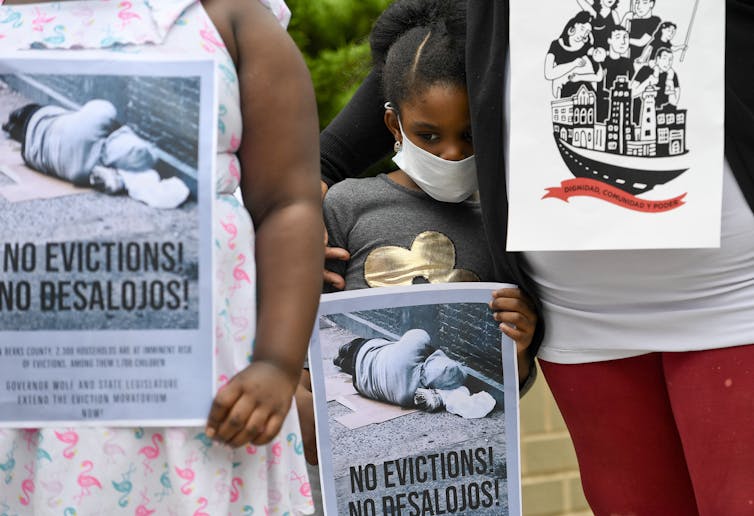 A young girl holds a bilingual sign reading 'no evictions/no desalojos' while being comforted by her sister