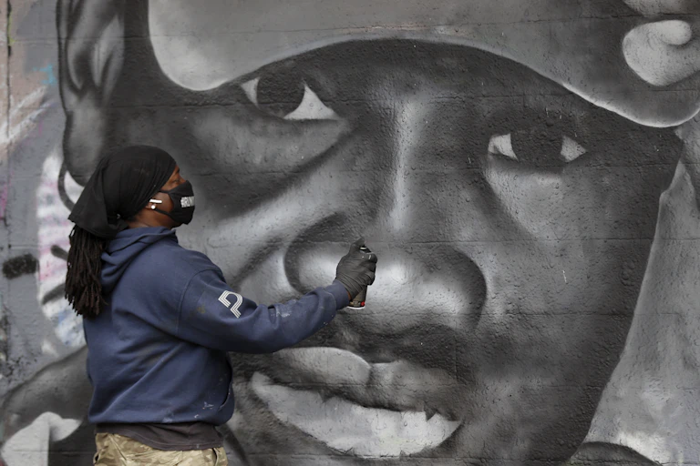 A man in a facemask holding a spraypaint can completes a mural paying tribute to Ahmaud Arbery.