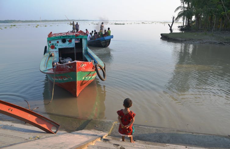 A young girl stands on a concrete bank as a red, wooden boat returns from fishing.