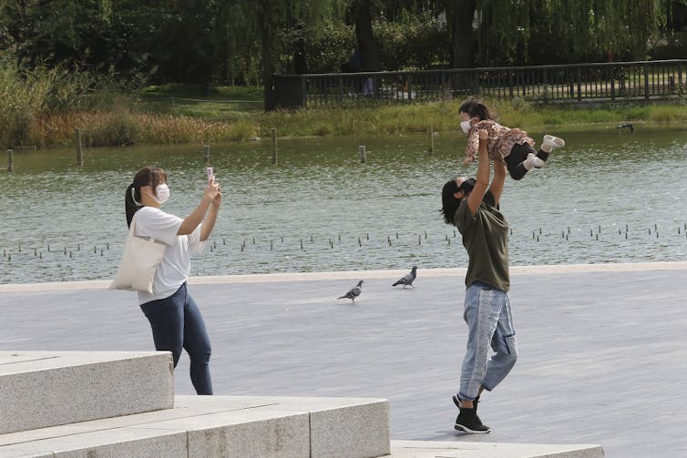 A person swings a baby up in the air as a woman takes a photo, all three wearing masks.