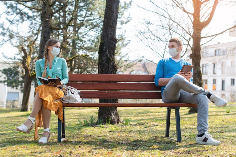 A man and a woman sit apart on a park bench wearing face masks.
