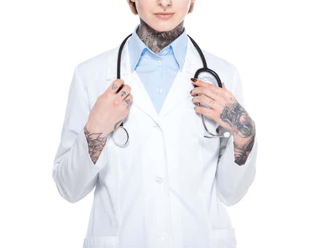 in a white medical jacket with tattoos on her neck and hands and a stethoscope around her neck