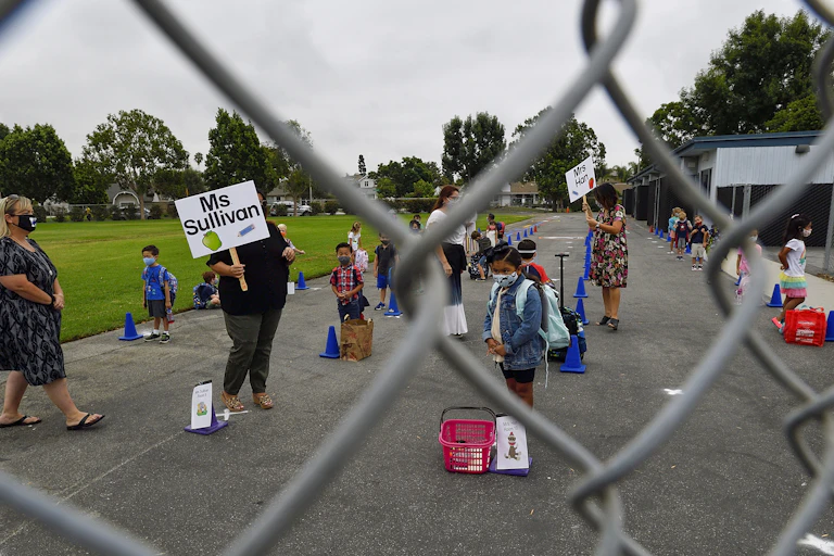 Teachers hold signs with their names as children gather outside their school wearing masks.