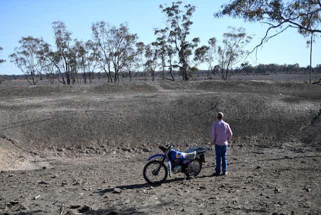 A man stands beside a motorbike, looking at a dry dam.