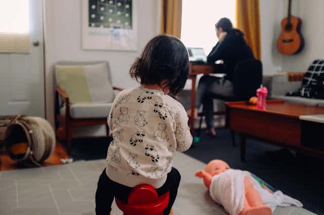 Woman working on her laptop in lounge room with a small child playing behind her