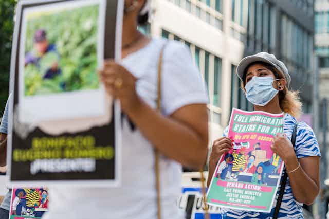 A woman wearing a mask carries a sign calling for full immigration status for seasonal workers.