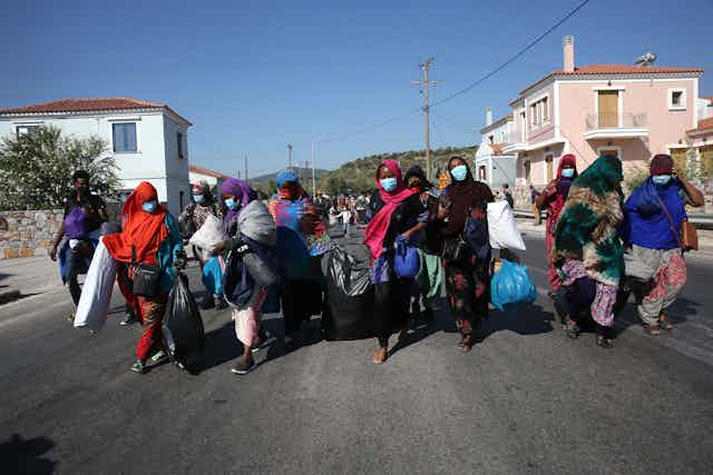 Women wearing headscarves and masks carry their belongings down a sunny street.