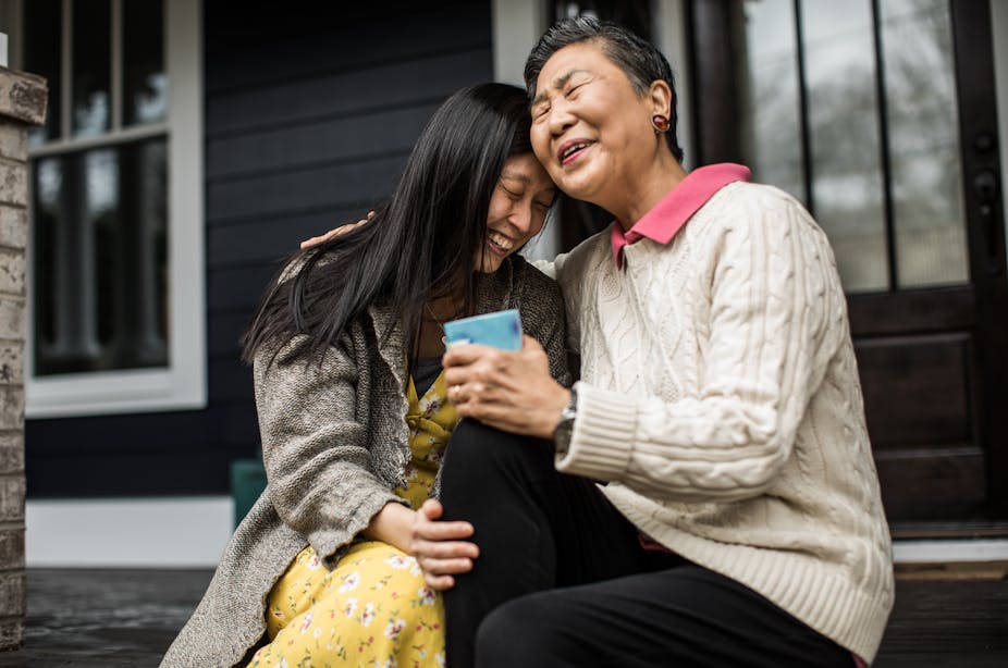 An Asian woman laughs with an elderly woman whas they're sitting onunder a porch.