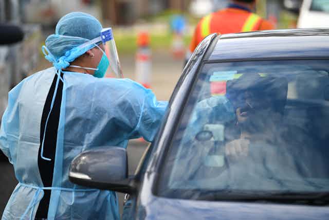 A woman in PPE performs a COVID test on a person in their car.