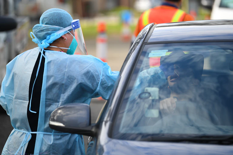 A woman in PPE performs a COVID test on a person in their car.