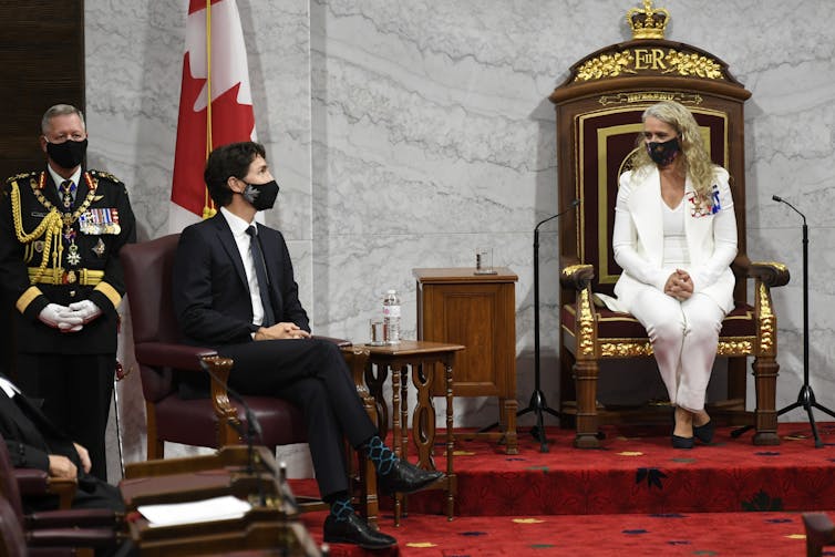 Gov. Gen. Julie Payette, wearing a white suit, sits in the ornate chair reserved for the governor general, on a raised platform. The prime minister sits in front of her on the left, with a man wearing a uniform behind him. They all wear black face masks.