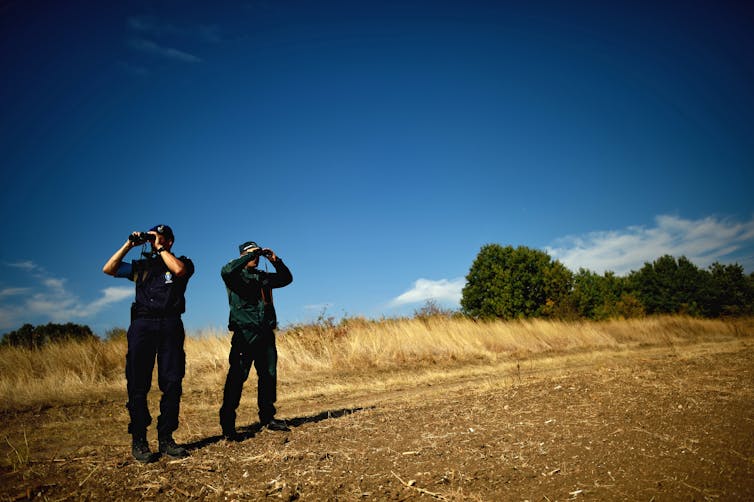 Two men in dark uniform look through binoculars.