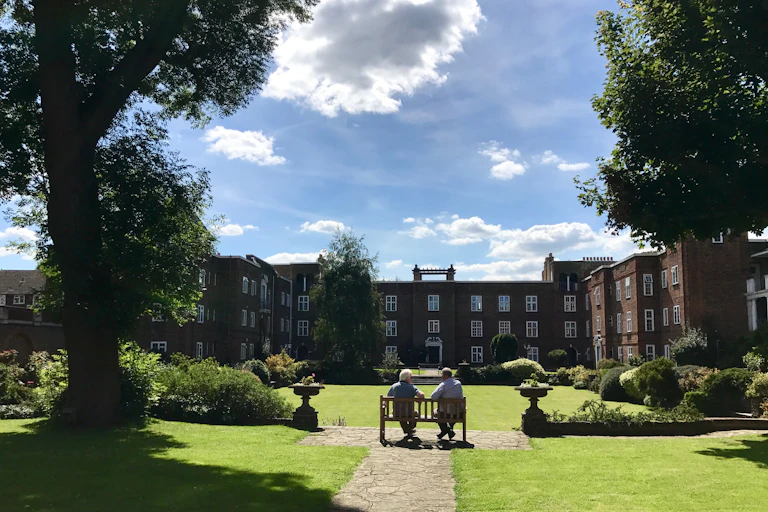 Two elderly sitting on a bench in the grounds of a nursing home on a sunny day.