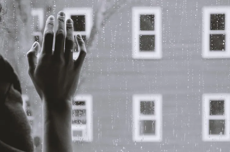 A woman's hand rests against a rain-spattered window pane.