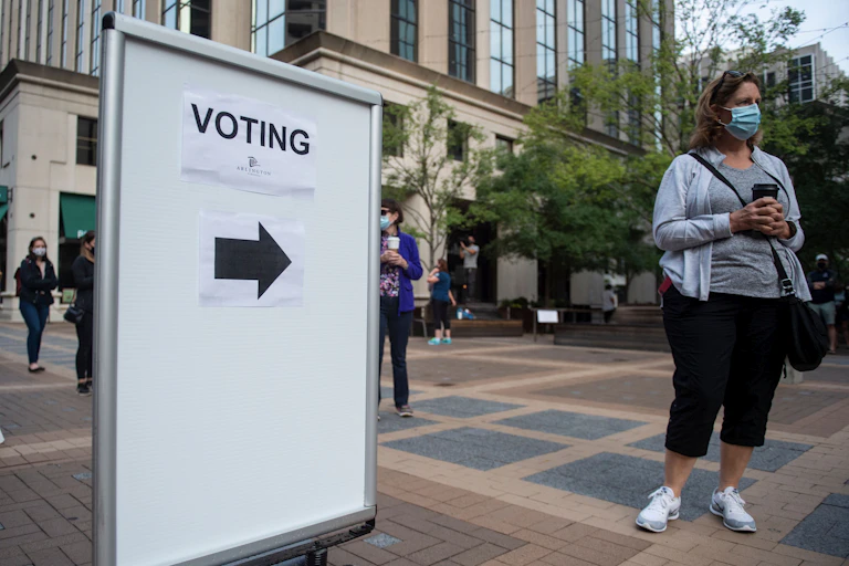 A woman with a mask stands next to a sign pointing to a voting station.