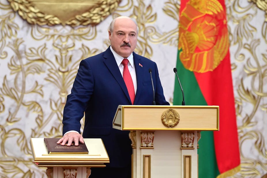 Lukashenko stands behind a lectern with his hand on a brown leather-bound book