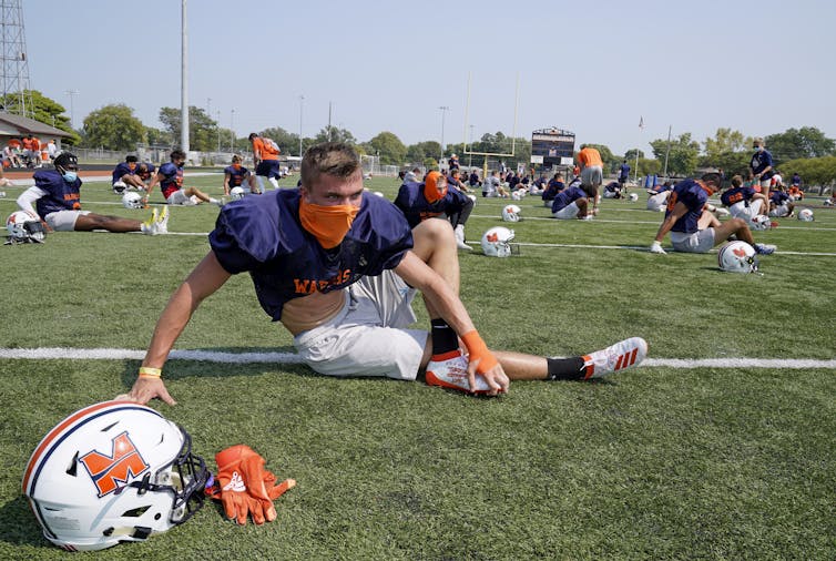 A college football player wearing a mask stretches during practice.