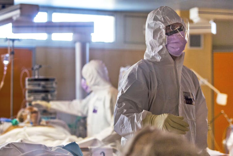 Two health-care workers completely covered by white protective suits and face masks tend to patients on gurneys.