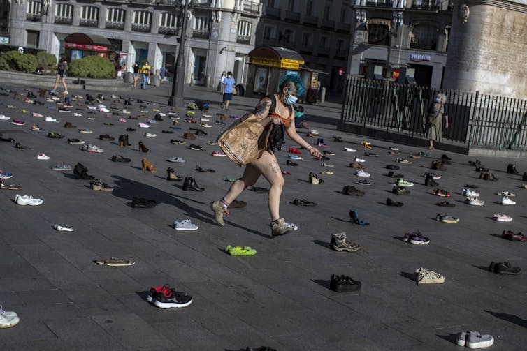 A woman with blue hair carries a paper bag and a pair of shoes as she steps among hundreds of pairs of shoes laid out in a grid in the public square.