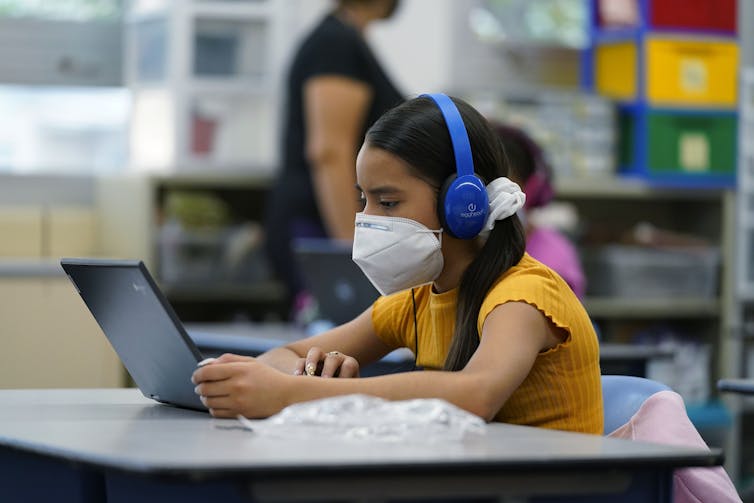 A middle school girl wearing blue headphones and a face mask looks at her laptop.