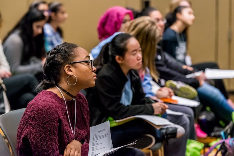 Young women sit in an audience listening to a speaker.