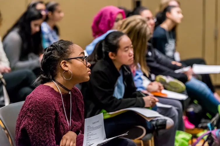 Young women sit in an audience listening to a speaker.