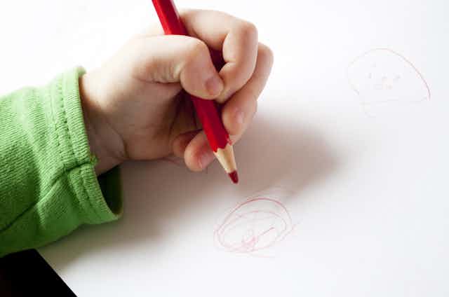 A child's left hand holding a pencila and drawing on a piece of paper.